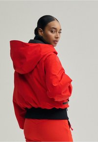 Piros kapucnis dzseki fekete béléssel, elasztikus derékpánttal és laza szabással. Simább anyag, cipzáras zseb részlettel.