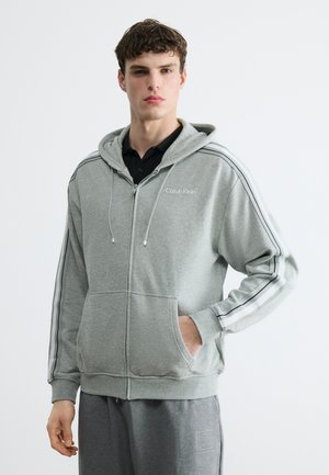 Νεαρός άνδρας που φοράει γκρι φούτερ με φερμουάρ και λευκές ρίγες στα μανίκια και το λογότυπο Calvin Klein, στέκεται με το ένα χέρι στην τσέπη.