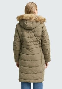 Kobieta w długim oliwkowym puchowym płaszczu zimowym z kapturem wykończonym futrem, widoczna z tyłu, w połączeniu z niebieskimi dżinsami.