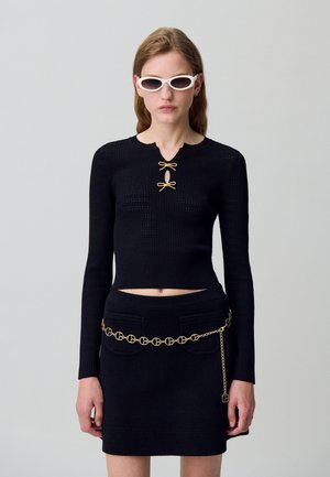 Crop top noir en tricot à manches longues avec des accents de nœud dorés et un décolleté en forme de clé ouvert ; associé à une mini jupe noire ornée d'une ceinture en chaîne dorée.
