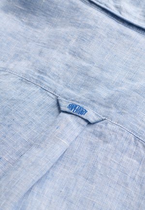 Hellblauer strukturierter Stoff mit einer Schlaufe am hinteren Kragen, mit einem kleinen blau bestickten Markennamen.