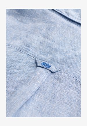 Light blue textured fabric with a loop tab sewn at the back collar, featuring a small blue embroidered brand name.