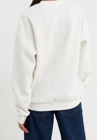 Felpa bianca oversize con spalle dropped, morbido al tatto e polsini a coste, abbinata a jeans scuri con cuciture in evidenza.