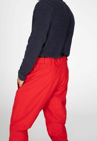 Personne portant un pantalon rouge vif et un haut à manches longues bleu marine foncé, une main dans la poche, sur un fond uni.