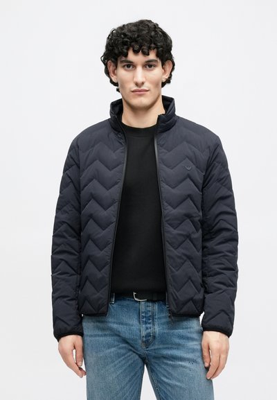 Tmavomodrá prešívaná vetrovka s vysokým golierom, so zipsovým zapínaním a vzorom šípok, v kombinácii s čiernou košeľou a svetlými džínsami z denimu.