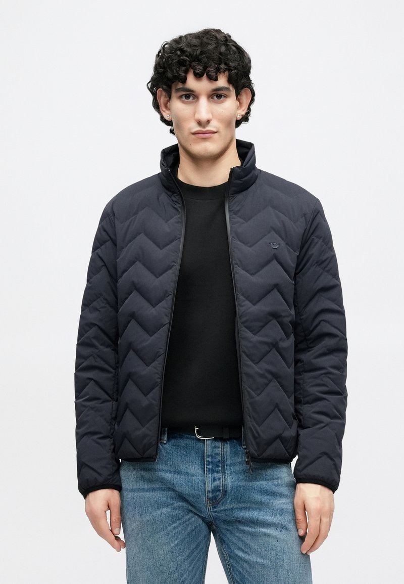 Veste matelassée bleu marine avec un col montant, dotée d'une fermeture éclair et d'un motif chevron, associée à une chemise noire et un jean en denim clair.