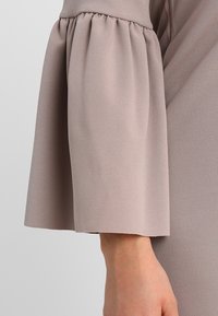 Blouse à manches longues en beige clair, fabriquée dans un tissu lisse et léger avec un détail de bouffant froncé au niveau du coude, présentant un poignet évasé.