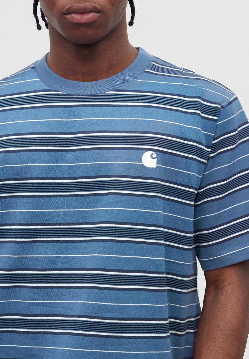 Junge Person mit geflochtenem Haar, die ein blau gestreiftes T-Shirt mit kleinem weißem Logo auf der linken Brust vor einem einfarbigen Hintergrund trägt.