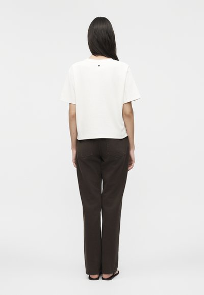 Γυναίκα με μακριά σκούρα μαλλιά, που φοράει λευκό μπλουζάκι με κοντά μανίκια και σκούρο καφέ παντελόνι, στέκεται πλάτη σε απλό φόντο.