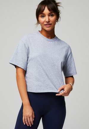 Junge Frau mit braunen Haaren zu einem Dutt gebunden, trägt ein hellgraues, bauchfreies T-Shirt und marineblaue Leggings, hält den Saum ihres Shirts vor einem einfarbigen Hintergrund fest.