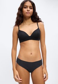 Donna con lunghi capelli ricci che indossa un reggiseno nero e mutandine bikini nere, in piedi davanti a uno sfondo bianco semplice.