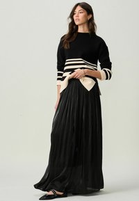 Pull rayé noir et crème à coupe décontractée associé à une jupe noire longue et plissée. La texture lisse et des chaussures noires pointues complètent le look.