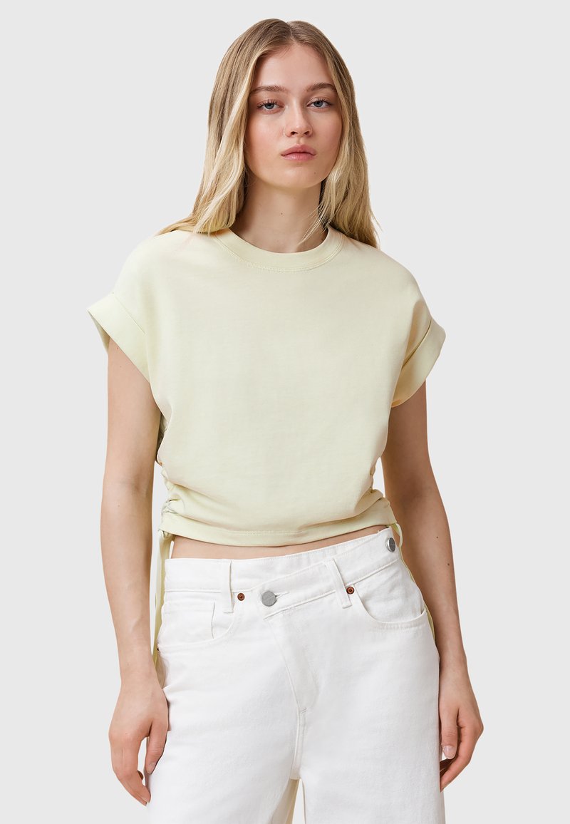 Lichtgele cropped t-shirt met opgerolde mouwen en zijsplitten, gecombineerd met witte high-waisted jeans met zilveren hardware en zakken.