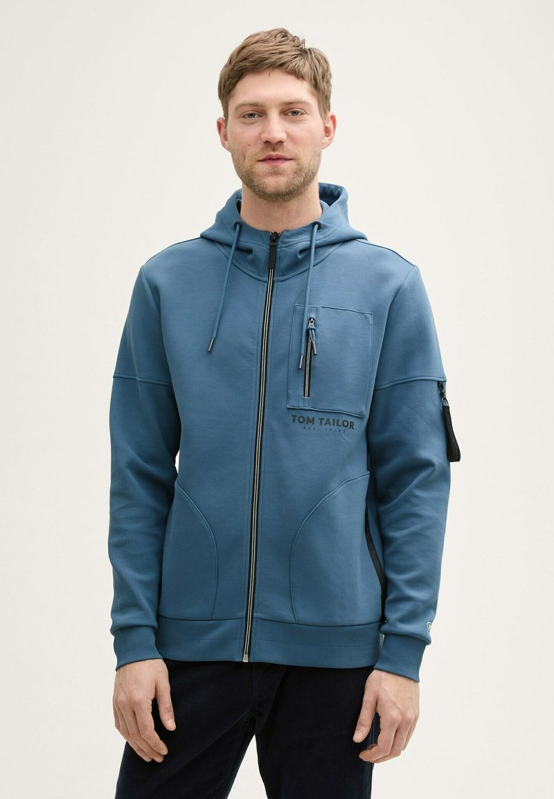 Niebieska, zapinana na zamek bluza z kapturem, o teksturowanej tkaninie, z bocznymi kieszeniami i kieszonką na piersi. Zawiera czarne logo i zamki błyskawiczne jako detale.