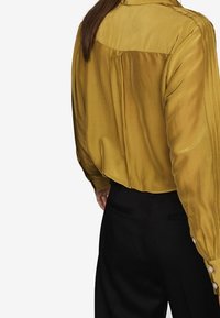 Blouse en satin jaune moutarde avec des plis aux épaules, des détails de boutons et une coupe décontractée, assortie à un pantalon noir ajusté.