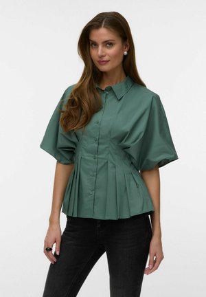 Femme aux longs cheveux bruns porte un chemisier plissé vert avec de larges manches courtes et un pantalon noir, debout devant un fond uni.