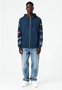Sweat à capuche zippé bleu marine avec des imprimés graphiques rouges, blancs et multicolores sur les manches et la poitrine. Porté avec un jean large bleu clair et des baskets blanches.