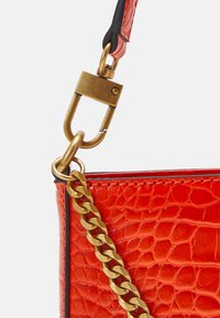 Gros plan sur un coin de sac rouge à texture crocodile avec une chaîne dorée en bandoulière et un fermoir rectangulaire en or fixé à une sangle en cuir noir et rouge.