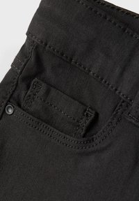 Tissu en denim noir avec un gros plan sur une poche arrière. Présente un petit overlay de poche avec des coutures visibles et un accent de bouton en métal.