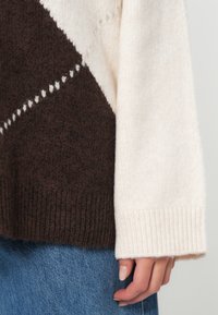 Χέρι resting δίπλα, φορώντας ένα κρεμ και καφέ πουλόβερ πλεκτό με διαγώνιο σχέδιο, συνδυασμένο με μπλε τζιν.