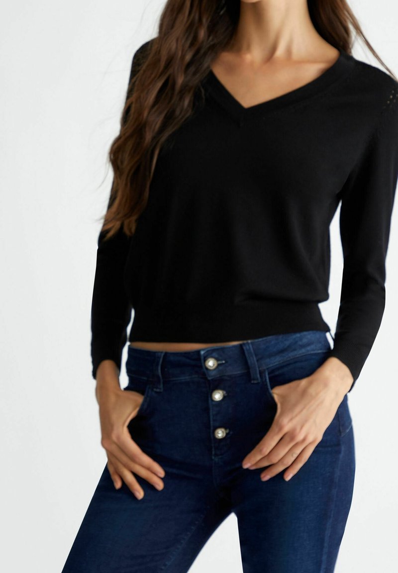 Schwarzer langärmeliger V-Ausschnitt-Pullover kombiniert mit dunklen Jeans im hohen Taillenstil und fünf silbernen Knopfakzenten. Glatte Textur.