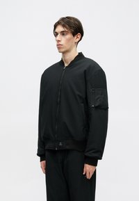 Μαύρο bomber μπουφάν με φερμουάρ μπροστά, ριμπ μανίκια και μέση. Διαθέτει μια παγκάκι τσέπη στον αριστερό μανίκι και λεία υφή.