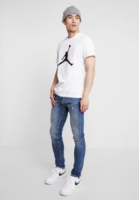 Hvid t-shirt med et sort Jordan-logo, parret med slidte blå jeans og hvide sneakers med et grønt tjek. Grå beanie på hovedet.