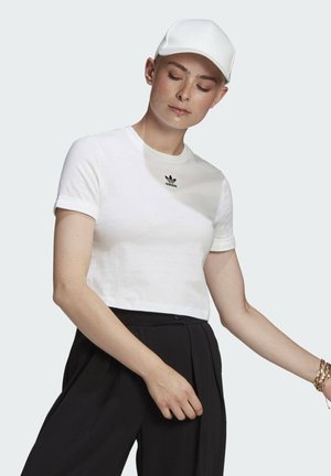 Biały skrócony t-shirt z krótkimi rękawami i czarnym logo Adidas na piersi, zestawiony z czarnymi spodniami z wysokim stanem. Biała czapka.