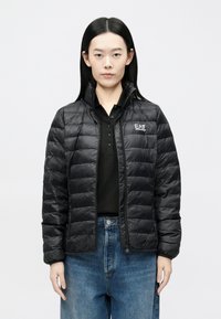 Melnā pūkau jaka ar horizontālām šuvēm, augstu apkakli, rāvējslēdzēja aizdari un logotipa detaļu. Valkāta virs melnas polo krekla, kombinēta ar zilām džinsa biksēm.