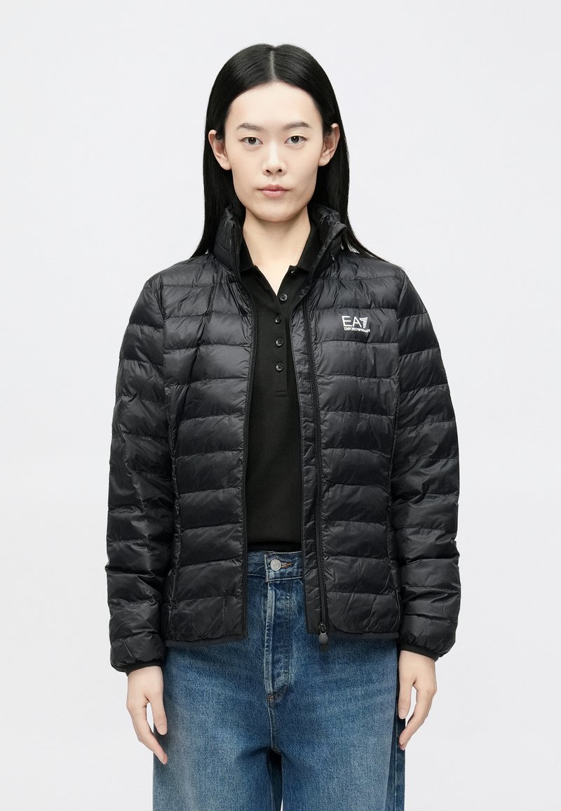 Melnā pūkau jaka ar horizontālām šuvēm, augstu apkakli, rāvējslēdzēja aizdari un logotipa detaļu. Valkāta virs melnas polo krekla, kombinēta ar zilām džinsa biksēm.