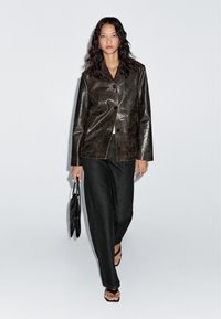 Vrouw met lang zwart haar, gekleed in een donkerbruine leren jas, zwarte wijde jeans, met een zwarte handtas en zwarte sandalen.