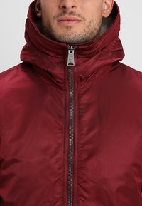 Jacket bordeaux com colarinho alto, apresentando um fecho éclair frontal e um tecido liso e brilhante. O design inclui um corte ajustado e detalhes de hardware mínimos.