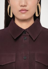 Cămașă bordo cu guler, închidere cu nasturi și două buzunare la piept. Prezintă cercei mari, ovali, texturati, de culoare aurie. Finisaj din material neted.