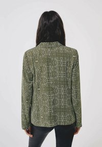 Grön mönstrad jacka med långa ärmar och krage. Designen inkluderar vita abstrakta former på ett texturerat tyg.