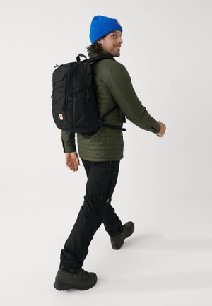 Zwarte rugzak met meerdere vakken, textiel met structuur, verstelbare banden, gedragen door een persoon in een groene jas en een blauwe beanie, die loopt.