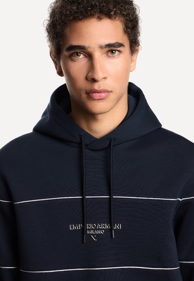 Giovane uomo con capelli ricci che indossa una felpa navy blue di Emporio Armani con strisce orizzontali bianche e testo del logo sul petto.
