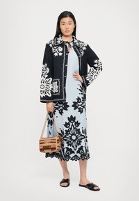 Jacke mit Schwarz-Weiß-Blumenmuster über einem hellblauen Kleid. Das Model hält eine mehrfarbige, geflochtene Tasche und trägt schwarze Slidesandalen.