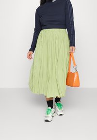 Kvinna i en marinblå Adidas långärmad tröja, ljusgrön plisserad midiklänning, vita sneakers med gröna snören och håller en orange handväska.