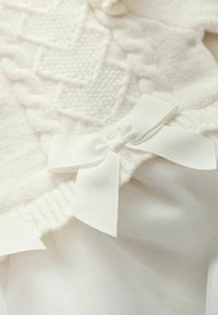 Vit plagg med texturerade mönster, en vävd design och en mjuk tygfinish. Inkluderar en dekorativ vit rosett.