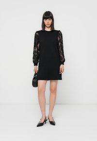 Woman wearing a black mini dress with sheer floral puff sleeves, black slingback heels, and holding a small black handbag, standing against white wall.
