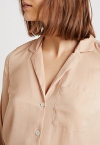 Light pink blouse with a button front, notch collar, and a single left chest pocket. Made of smooth, lightweight fabric.