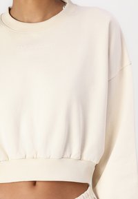 Kremfarget cropped sweatshirt med ribbede mansjetter og nederkant. Har brodert merkenavn "CARLO COLUCCI" i en subtil tone på forsiden.