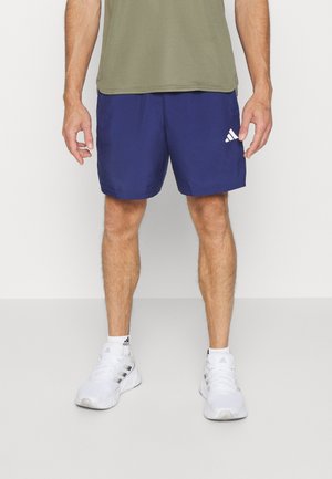 Man draagt marineblauwe Adidas-shorts, olijfgroen shirt, witte Adidas-sokken en witte Adidas-sneakers, staand tegen een effen achtergrond.