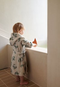 Šviesiai pilkas chalatas su žaliomis krabų raštais, surištas juosmens srityje, vaikas laiko oranžinę popierinę valtį šalia seklos, šviesiai žalios vandens baseino.