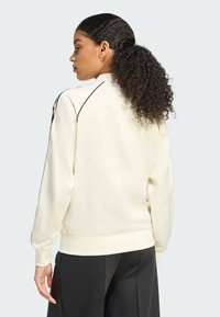 Sudadera crema con cuello alto, puños acanalados y acentos negros. Textura suave, corte relajado y diseño de costura en los hombros distintivo.