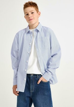 En prickig skjorta i blått och vitt med lång ärm, bröstficka; buren över en vit t-shirt och tillsammans med mörkblå jeans.