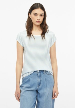 Frau mit langen braunen Haaren trägt ein hellblau-weiß gestreiftes kurzärmliges Shirt und lockere hellblaue Jeans, steht vor weißem Hintergrund.