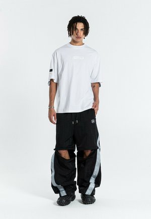 Joven de pie mirando hacia adelante con una camiseta blanca extragrande y pantalones negros con piernas inferiores desmontables y rayas laterales grises.