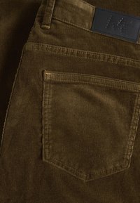 Brown corduroy pants feature a back pocket, subtle ribbed texture, and yellow stitching accents. A black leather brand tag is visible.
