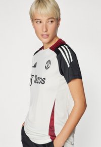 Balta sportinė marškinė su juodais ir burgundiškais akcentais, su trumpomis rankovėmis, Adidas juostomis ir „Manchester United“ logotipu.